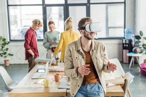 Preview: smiling male designer gesturing with hands while having virtual reality experience in loft office