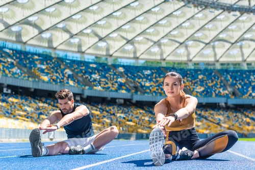 Preview: young sportive male and female joggers sitting on running track and stretching at sports stadium