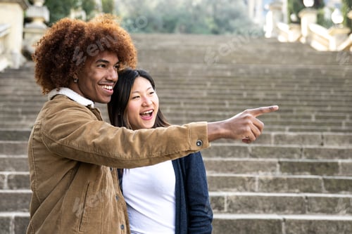 Preview: Lovely multiracial couple pointing to the right in a park.Two happy diverse young persons smiling