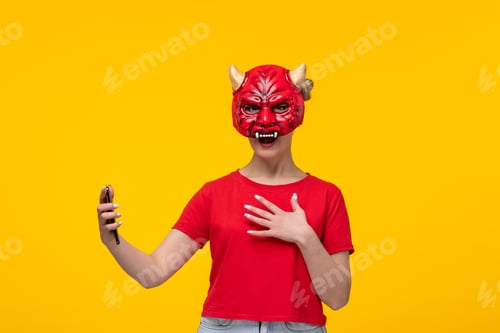 Preview: young female wearing scary devil mask using her phone yellow background ugly