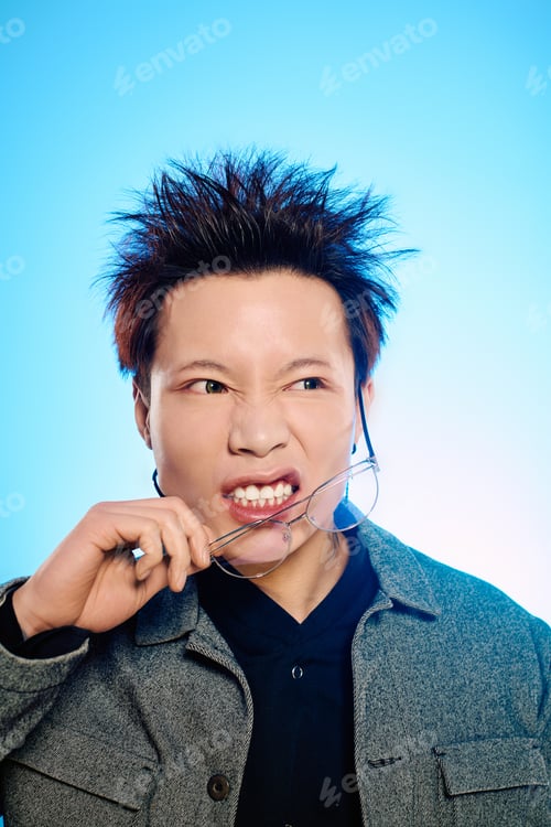 Preview: Stylish young asian man playfully bites glasses in a creative studio setting under vibrant lighting
