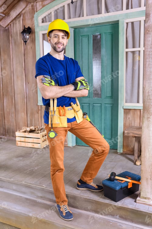 Preview: smiling carpenter in helmet with building tools standing on porch and looking to camera