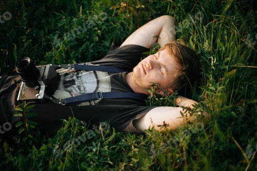 Preview: Stylish hipster man with film photo camera lying in green grass and relaxing in sunny meadow
