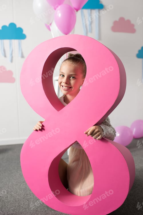 Preview: adorable little girl holding big pink decorative number eight and smiling at camera at birthday