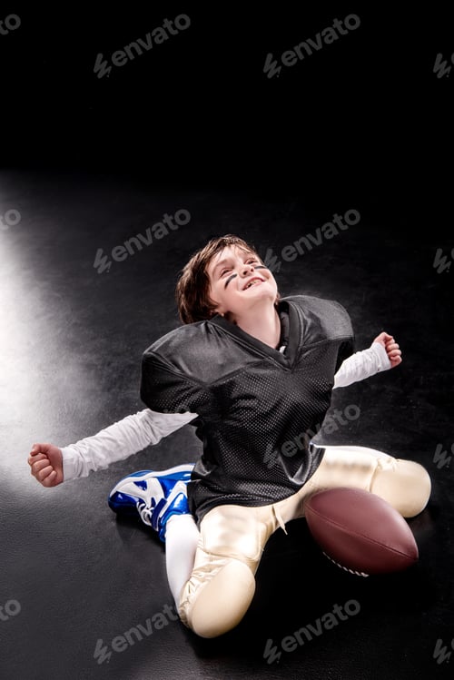 Preview: Cheerful boy american football player sitting with ball and triumphing