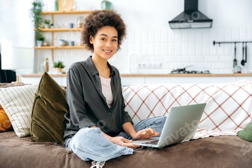 Preview: Lovely glad african american woman with short curly hair, in stylish wear, sit on a sofa in living