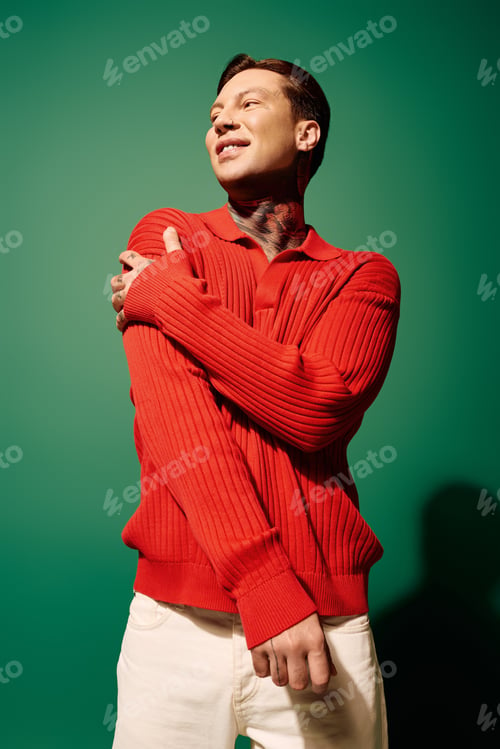 Preview: Young man with tattoos poses confidently against a vibrant green backdrop wearing a red sweater