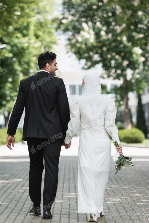 Preview: happy groom holding hands with muslim woman with wedding bouquet and walking together