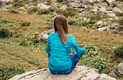 Preview: Rear view of Meditating young woman in blue sportswear sitting on a stone mountain valley