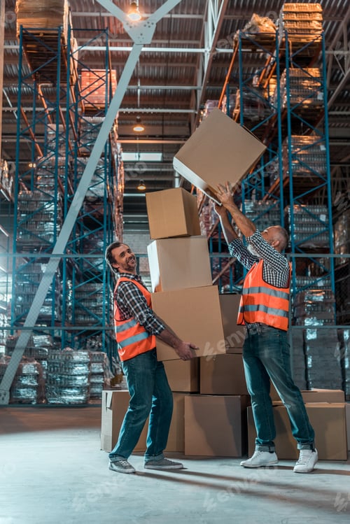 Preview: full length view of two workers in vests holding boxes in warehouse