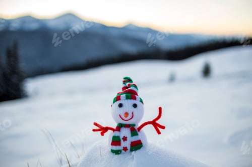 Preview: Small funny toy baby snowman in knitted hat and scarf in deep snow outdoor on blurred snow covered