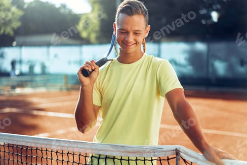 Preview: Young man teenager with tennis racket standing near net on clay court