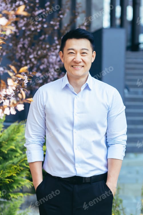 Preview: Vertical frame, Asian man in white shirt looks at camera and smiles, hands in pockets