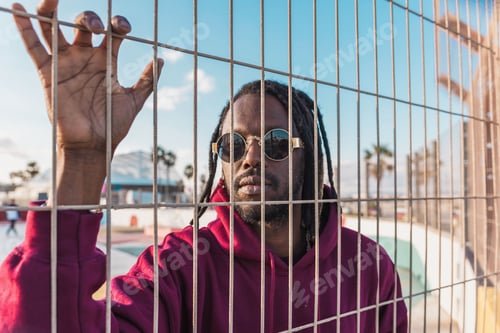 Preview: Black man with dreadlocks and sunglasses holding a metal fence with his hand