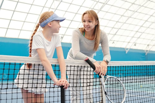 Preview: Cute girl in white sportswear and her trainer with tennis rackets talking by net