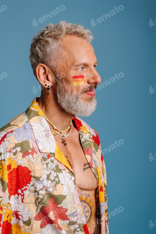 Preview: Mature gay man with rainbow flag make-up looking away while standing against blue background