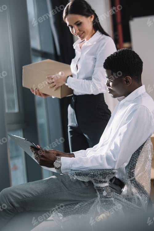 Preview: multiethnic businessman and businesswoman looking at clipboard while relocating in new office