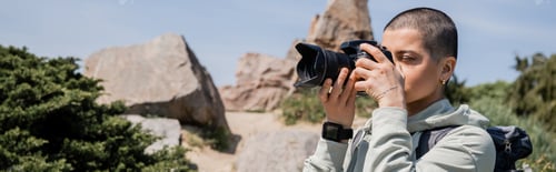 Preview: Young tattooed and short haired woman hiker with backpack taking photo on digital camera