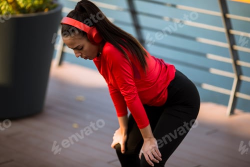 Preview: Young sporty woman with earphones taking break after a run in the urban environment