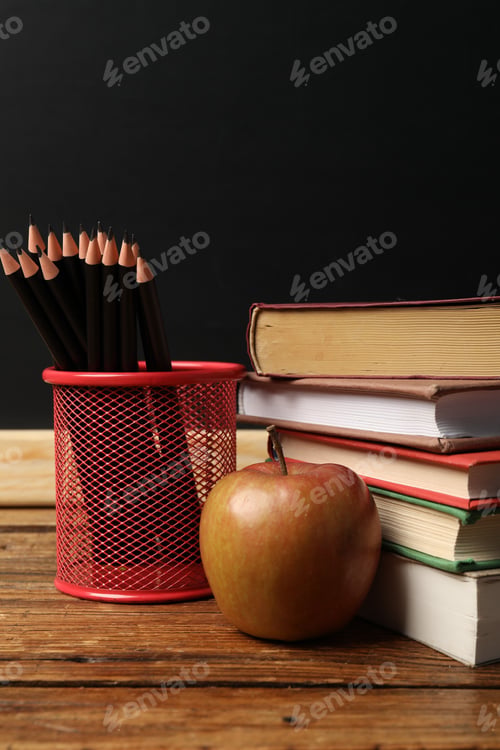 Preview: Different books, pencils and apple on wooden table near blackboard, closeup