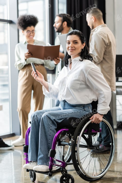 Preview: happy disabled businesswoman in wheelchair looking at camera near blurred multiethnic colleagues