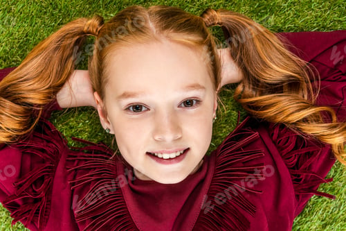 Preview: top view of cute schoolgirl with hands on head lying on lawn and looking at camera