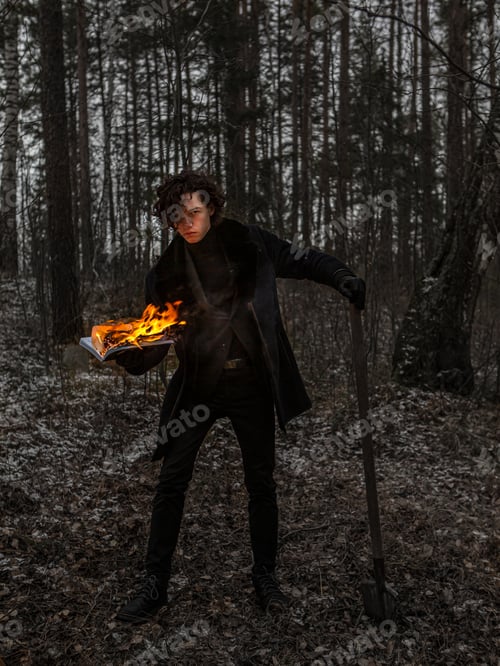 Preview: A Caucasian guy, a teenager, with long curly hair stands in the forest with a burning book