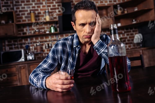 Preview: upset man sitting at table with bottle and glass of alcohol at home