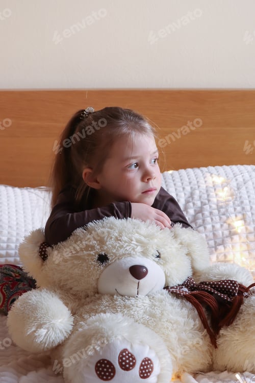 Preview: A little pretty girl looks with a bored look. A teddy bear sits nearby. Bored mood.