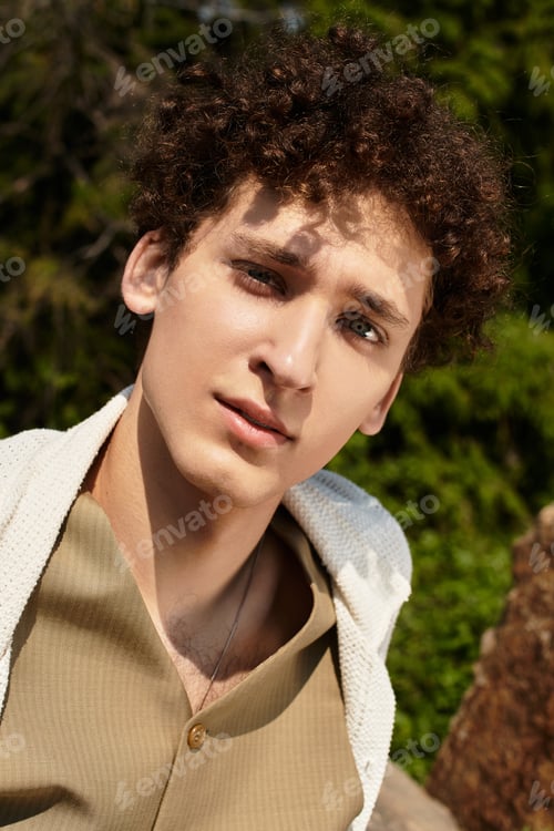 Preview: Curly-haired young man embraces summer fashion in a vibrant green park