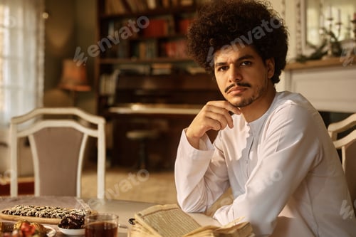 Preview: Young Arab man reading from Quran at dining table and looking at camera.
