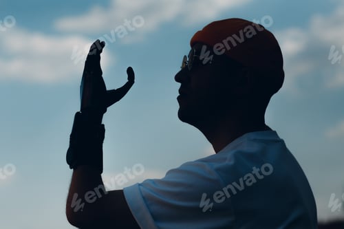 Preview: Young disabled man with artificial prosthetic hand in casual clothes silhouette against sky.