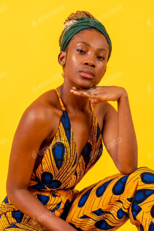 Preview: Young african woman with traditional costume on yellow background, sitting looking at camera