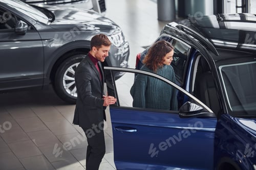 Preview: Professional salesman assisting young girl by choosing new modern automobile indoors