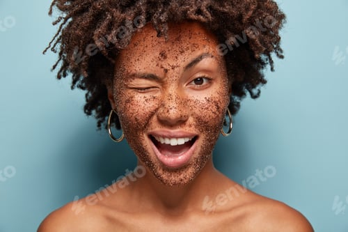 Preview: Image of pleased young curly woman blinks eye, has positive expression, applies black coffee mask, d