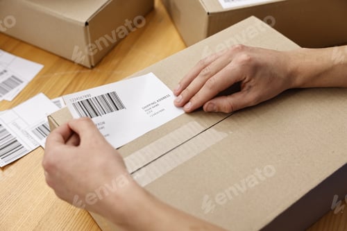 Preview: Man sticking shipping label with barcode on parcel at wooden table indoors, closeup