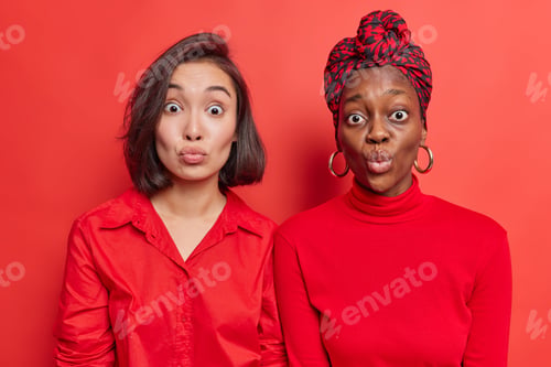 Preview: Diverse women best friends stand closely to each other keep lips rounded wait for kiss wears red clo