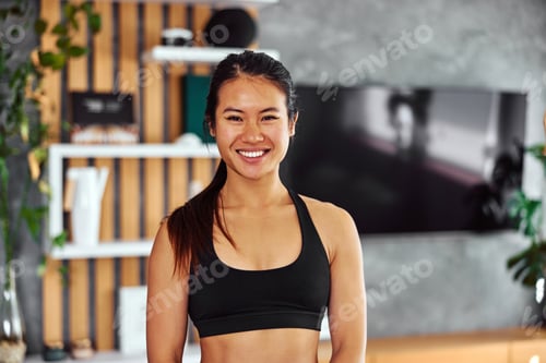 Preview: Portrait of a happy Asian female in a sports set, standing in the living room.