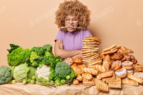 Preview: Displeased curly haired woman poses with arms folded asparagus in mouth feels temptation to eat
