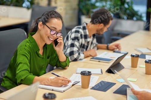 Preview: Young cheerful asian woman talking by phone and making some notes while working in the modern office