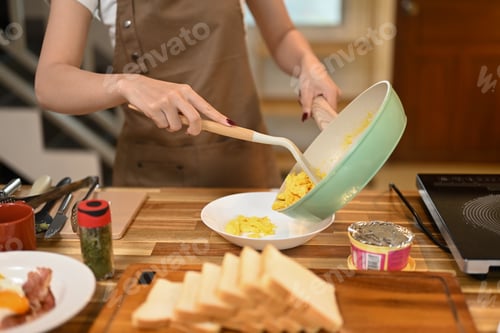 Preview: Young woman in brown apron serving scrambled eggs from a pan onto a white plate