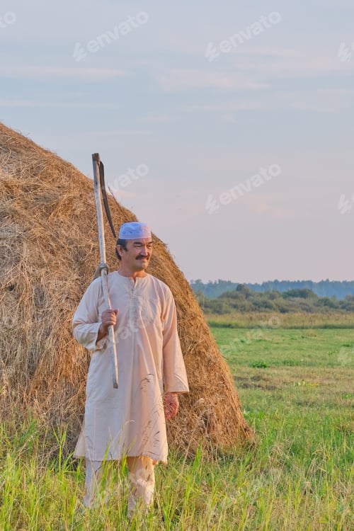 Preview: Senior Muslim man in traditional clothes leaves with hand scythe from hay field at sunset