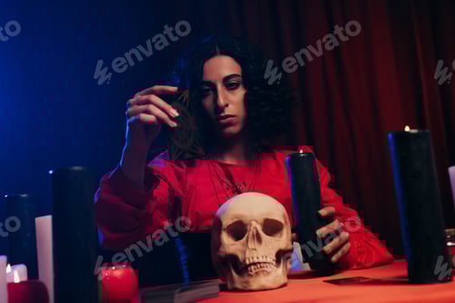 Preview: young medium sitting near skull and burning candles on dark background with red drapery