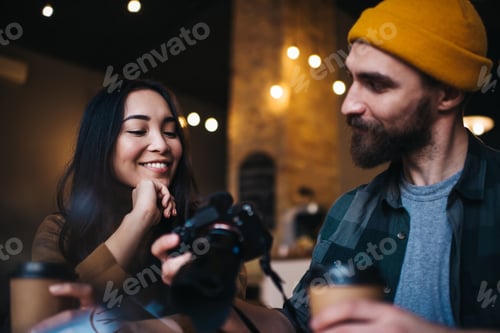 Preview: Positive multiracial couple viewing photos on camera while drinking coffee in cafe