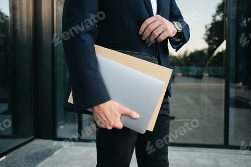 Preview: Close up man in classic black suit standing outdoor holding laptop and big envelope in hand