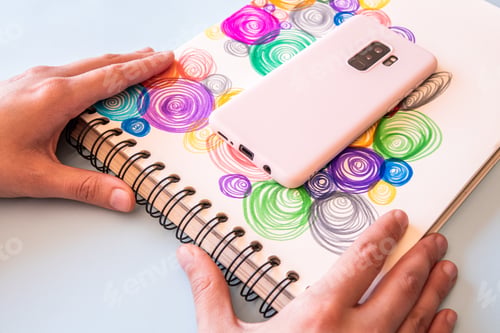 Preview: Closeup shot of a man's hands holding a notebook of drawn colorful spirals and a phone placed on it