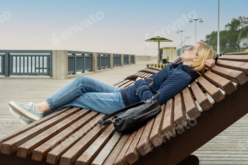 Preview: Young lonely woman on empty city embankment rests with her eyes closed on sun lounger