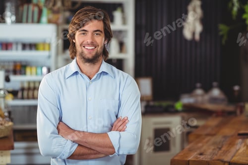 Preview: Bearded man standing with arms crossed in cafe with wooden service counter and glass cake domes