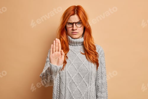 Preview: Angry irritated woman with red hair makes stop gesture stretches arm raises eyebrows and looks with