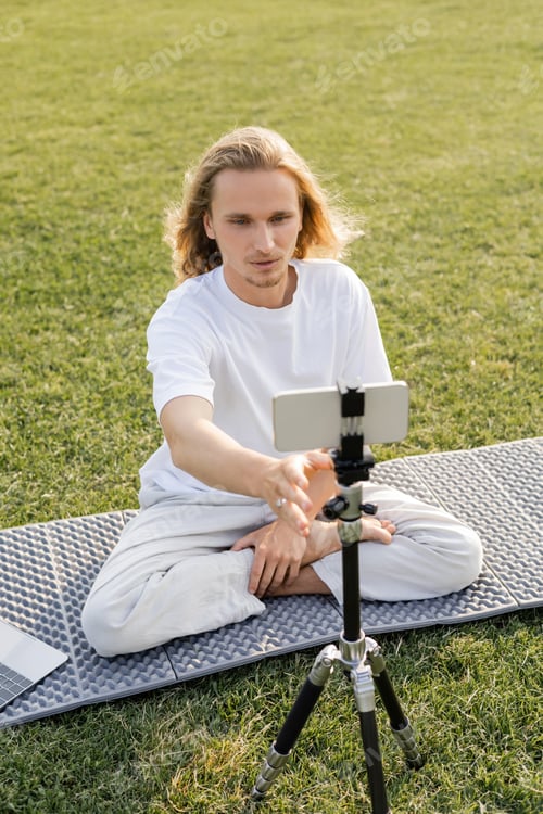 Preview: young yoga vlogger adjusting tripod with mobile phone while sitting in easy pose on grassy field